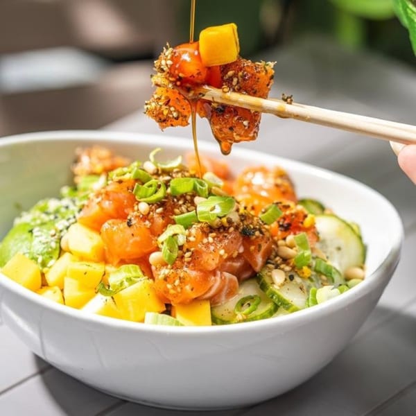 Chopsticks lifting a piece of salmon and mango from a healthy poke bowl with sauce drizzling.