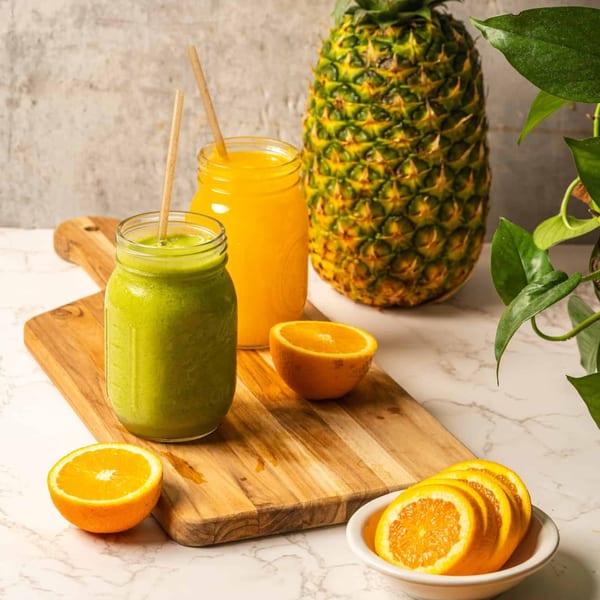 Two healthy smoothies, one green and one orange, with fresh pineapple and oranges on a wooden board.