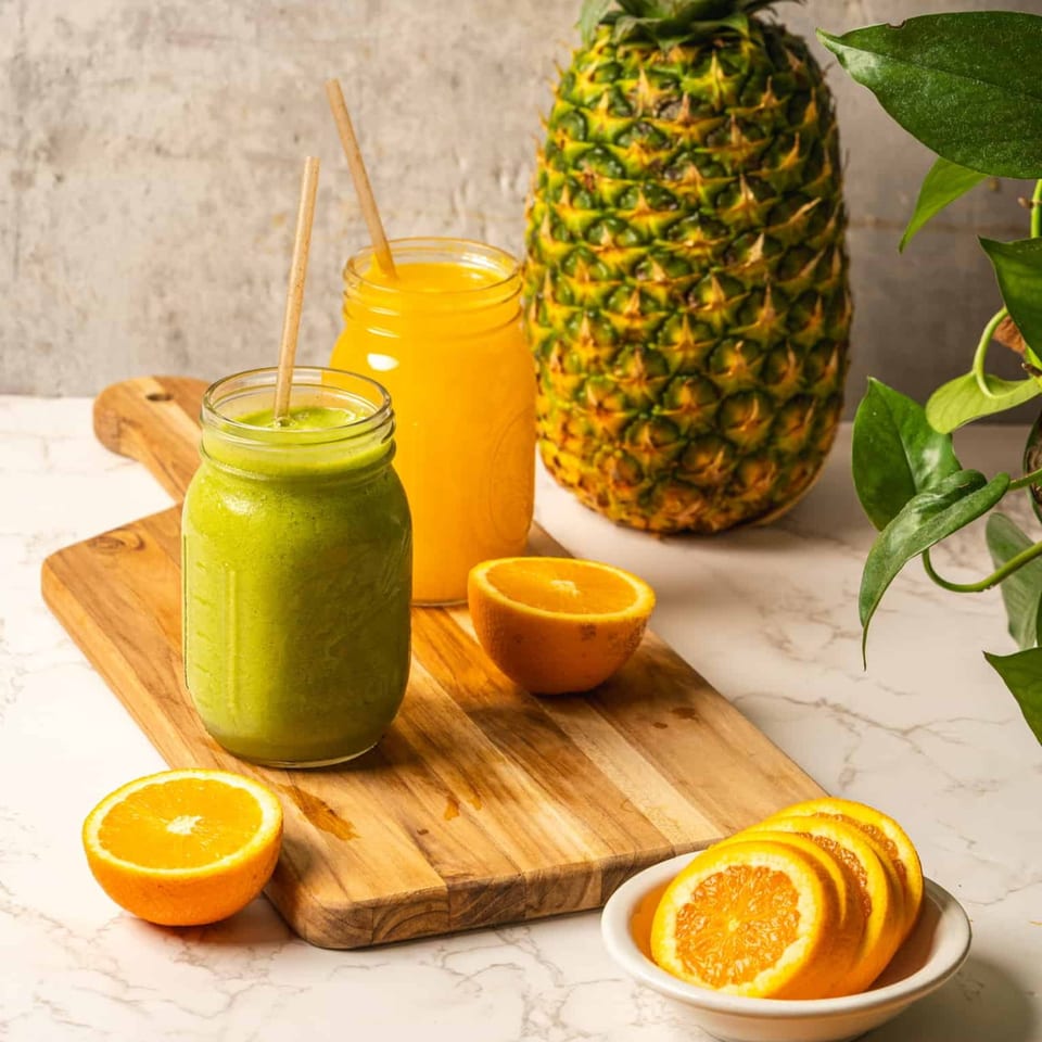 Two healthy smoothies, one green and one orange, with fresh pineapple and oranges on a wooden board.