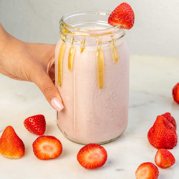 Hand holding a strawberry smoothie in a mason jar, drizzled with honey, surrounded by fresh strawberries.