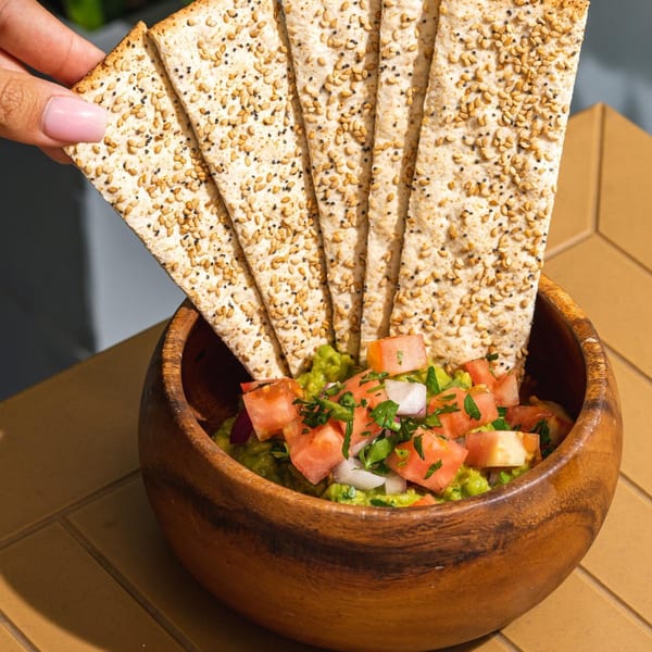 A hand holds a sesame and poppy seed flatbread cracker above a wooden bowl of guacamole with tomatoes and onions.