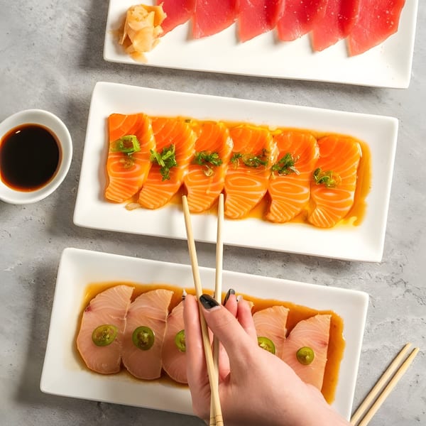 Overhead view of three plates of fresh sashimi, including salmon, tuna, and yellowtail, with chopsticks.