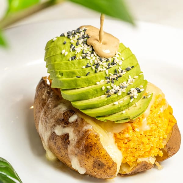 A baked potato topped with melted cheese, orange filling, sliced avocado, sesame seeds, and sauce.