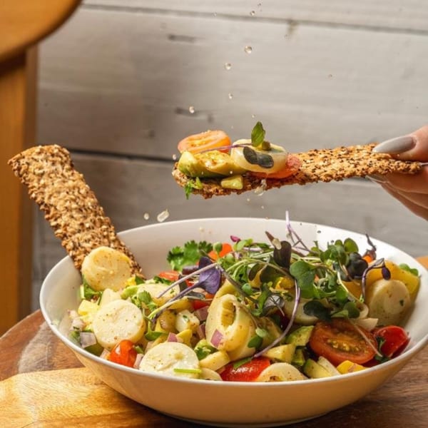 A hand holds a cracker topped with avocado and tomato over a fresh salad bowl.