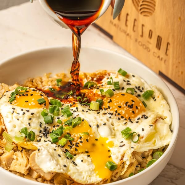 Soy sauce pouring over a bowl of fried eggs, rice, chicken, green onions, and sesame seeds.