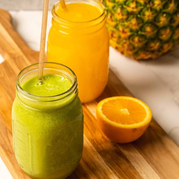 Two mason jars of green and orange smoothies, an orange half, and a pineapple on a wooden board.