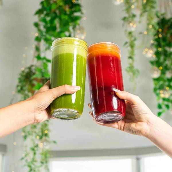 Two hands holding up tall glasses of vibrant green and red healthy juices, cheering.