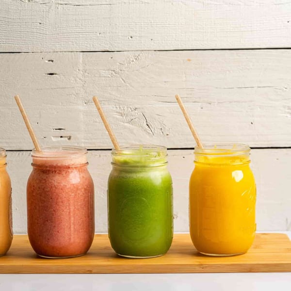 Three colorful smoothies in mason jars with straws on a wooden board.