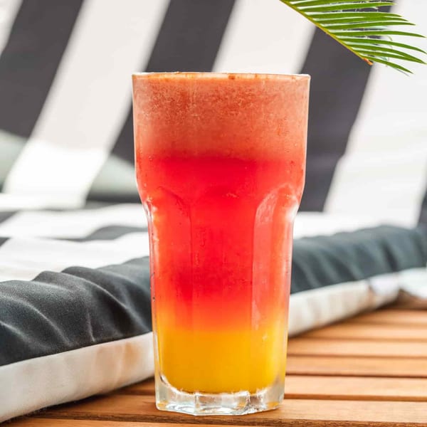 A vibrant, layered smoothie in a tall glass, with red, orange, and yellow colors, on a wooden table.