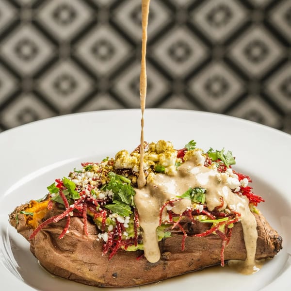 Sauce being drizzled over a loaded baked sweet potato with avocado, cheese, and red beet strings.
