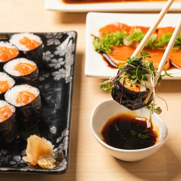 Chopsticks dipping a fresh sushi roll with microgreens into a bowl of soy sauce.
